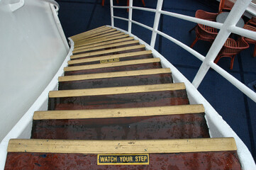 A high angle view shows wet wooden steps with watch your step warning signs on a curved staircase of a cruise ship deck near chairs. © nahhan