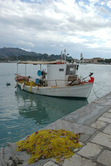 Fototapeta premium A traditional white wooden fishing boat moored at a stone dock with a pile of yellow fishing nets in Zakynthos port under a cloudy sky
