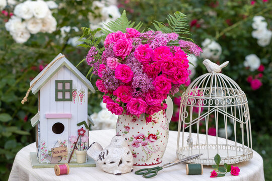 Arrangement mit Blumenstrau&szlig; mit pink Zwergrosen "Roxy" und Sommerspiere-Bl&uuml;ten, Vogelk&auml;fig und Vogelhaus im Sommergarten