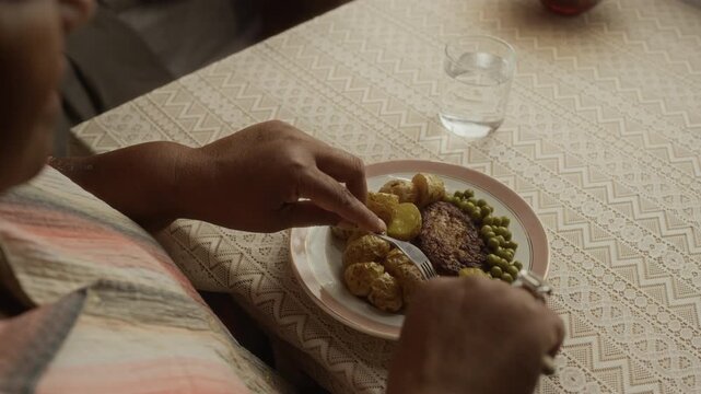 Over shoulder of senior African American woman eating homemade meal of potatoes, peas, and cutlet at dining table. Clip is suitable for senior care, healthy nutrition, and independent living campaigns