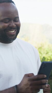 Vertical video: Smiling man reading phone on patio, sipping mug to relax, in white tee and bracelet