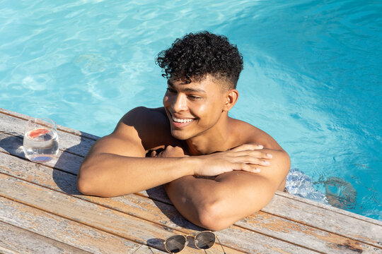 Shirtless man smiling, leaning on wooden deck by pool with round sunglasses, citrus glass