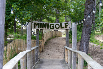Obraz premium Wooden bridge leading to a minigolf course entrance surrounded by trees and a sandy path, with decorative lights strung overhead for ambiance