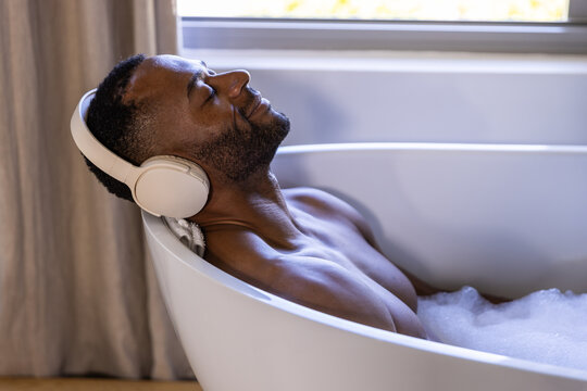Mature shirtless African man reclining in bathroom tub listening with headphones, towel behind head