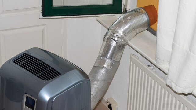 A portable monoblock air conditioner in a room with its flexible exhaust hose wrapped in thermal insulation tape and foil, vented outside through a sealed open window to prevent hot air from entering.