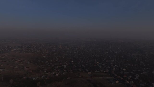 A high altitude aerial panorama of a scenic Armenian town split by a deep rocky canyon, showcasing houses, autumn trees, and distant misty mountains under a hazy sky.

