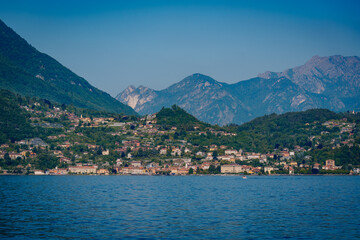 Fototapeta premium Scenic view of a lakeside village nestled among green hills and mountains under a clear blue sky, reflecting on the Menaggio