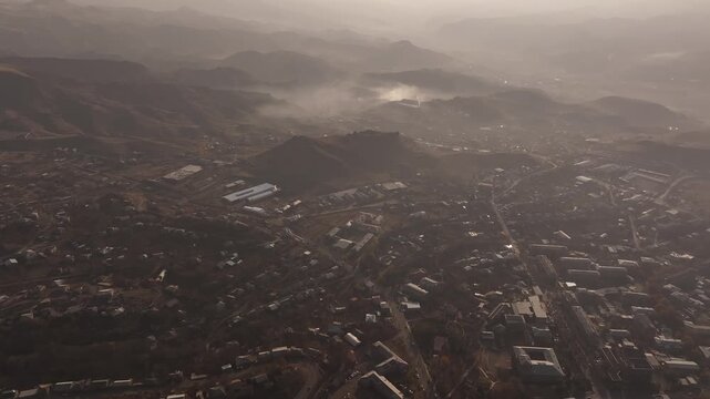 A high altitude aerial panorama of a scenic Armenian town split by a deep rocky canyon, showcasing houses, autumn trees, and distant misty mountains under a hazy sky.

