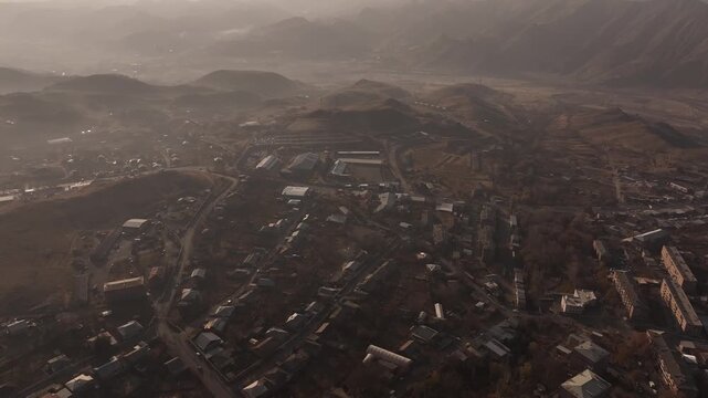 A high altitude aerial panorama of a scenic Armenian town split by a deep rocky canyon, showcasing houses, autumn trees, and distant misty mountains under a hazy sky.

