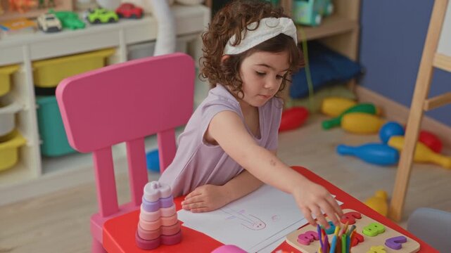 Girl drawing on paper with pencil, hand resting on page at a small colorful table in a studio; creative concentration.