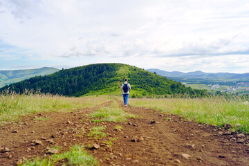Obraz premium Small figure walking through wide upland scenery with winding route stretching toward forested hills in the distance. Conveys landscape-scale recreation and exploration of expansive environments.