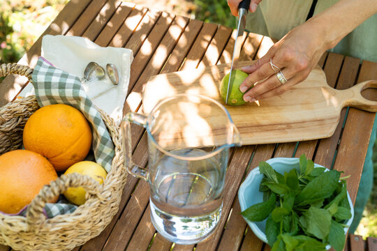 Woman using a knife to slice a fresh lime on a wooden board to make a homemade summer cocktail. Summer party