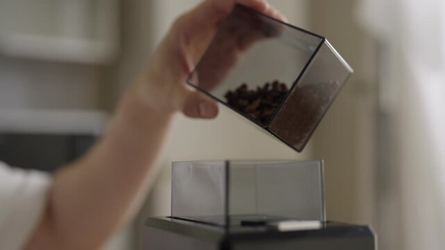 Unrecognizable person pouring whole roasted coffee beans from container into hopper of electric grinder, preparing to make fresh and aromatic cup of coffee, close-up. Shooting in slow motion