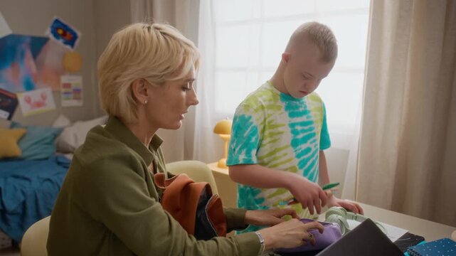 Middle-aged Caucasian woman helping Caucasian boy with Down syndrome pack school backpack. Footage perfect for promoting inclusive education and special needs support services