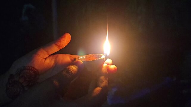 Woman's hand lighting a traditional oil lamp (Diya) during a Hindu prayer ceremony at night. The warm flame illuminates the hands adorned with mehndi and traditional jewelry