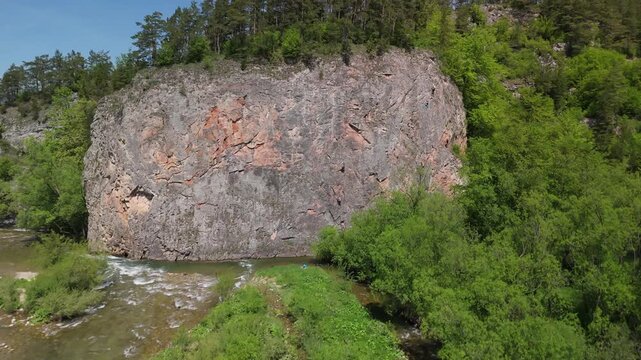 Aerial drone footage approaching a rocky cliff with athletes climbing, opening a view of a fast mountain river and green forest valley below.