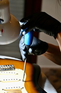 Vertical macro shot of a luthier servicing potentiometers and electronics on a sunburst stratocaster body under a lamp.