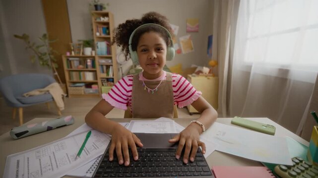 Portrait of African American child girl wearing headphones, talking, gesturing, typing on laptop during online class. Footage ideal for e-learning platforms and remote education commercials