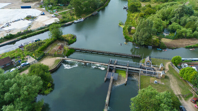 Aerial view of Feildes Weir Lock on the River Lee, featuring lock gates, flowing water, lush trees, and people kayaking on the river in Hoddesdon, England, United Kingdom.