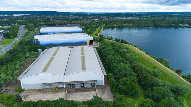 Aerial view of industrial factories adjacent to Willen Lake with large warehouses, loading bays, and surrounding green parkland in Milton Keynes, England, United Kingdom.