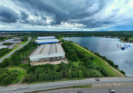 Aerial view of industrial factories adjacent to Willen Lake, surrounded by lush green trees and a road, under a dramatic cloudy sky in Milton Keynes, England, United Kingdom.