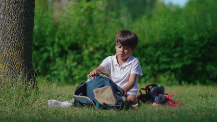 Naklejka premium Child placing soccer ball into backpack while sitting on grass at park capturing preparation departure routine and quiet moment of transition after outdoor play