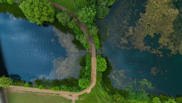 Aerial view of Willen Lake featuring a pedestrian path crossing over the water, surrounded by lush green trees and parkland in Milton Keynes, England, United Kingdom.