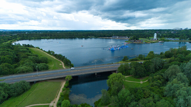 Aerial view of Willen Lake featuring a water park with inflatable obstacles, a bridge, and lush green trees under a cloudy sky in Milton Keynes, England, United Kingdom.