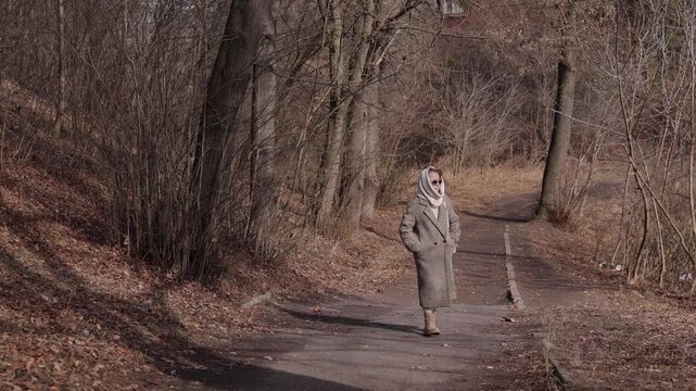 Senior lady wearing warm coat with woolen head-wrap walks along road among bare trees. Calm old woman strolls looking around in sunny autumn garden. Lifestyle of retirement