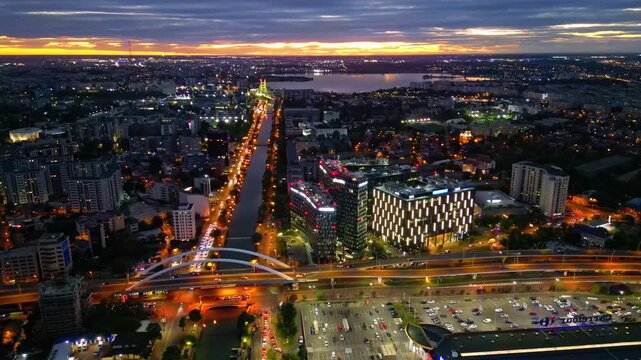 Drone aerial footage of a spectacular sunset in Bucharest, Romania. The camera captures the scenic Dambovita River in the Orhideea business area, showcasing modern office buildings, vibrant golden hou
