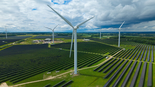 Aerial view of Hybrid Solar and Wind energy generation featuring rows of solar panels and wind turbines in a green field under a cloudy sky in Chelveston, England, United Kingdom.