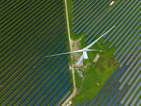 Aerial view of a wind turbine integrated within a large solar farm, showcasing hybrid renewable energy generation with rows of photovoltaic panels in Chelveston, England, United Kingdom.