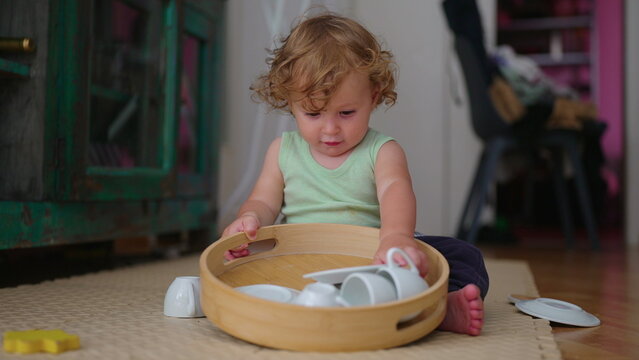Toddler sitting on floor sorting small cups and plates from wooden tray focused childhood moment of exploration organization and tactile play with household objects at home