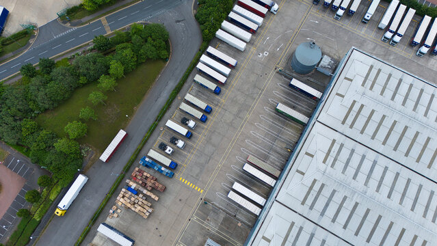 Aerial view of industrial warehouses with rows of parked semi-trailers and trucks in a loading yard, surrounded by green trees and paved roads in Kettering, England, United Kingdom.
