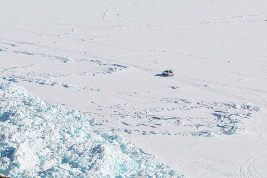 Aerial view of a car driving across the vast frozen surface of Lake Baikal with jagged ice ridges and snow in Baikalskoye, Buryatia, Russia.