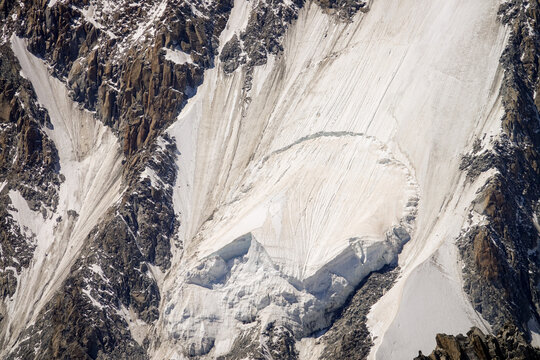 Aerial view of the glacier and rocky slopes near Punta Helbronner in the Mont Blanc massif, featuring deep crevasses and rugged alpine terrain in Courmayeur, Aosta Valley, Italy.