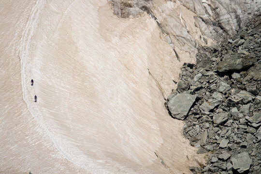 Aerial view of two hikers trekking across a glacier near rocky moraine slopes with snow and ice textures in Courmayeur, Aosta Valley, Italy.