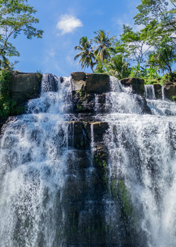 Aerial view of Cigangsa Waterfall cascading over layered rock formations surrounded by lush tropical palm trees under a bright blue sky in West Java, Indonesia.