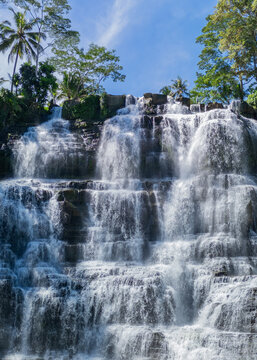 Aerial view of Cigangsa Waterfall cascading over tiered rock formations surrounded by lush tropical trees and dense foliage in West Java, Indonesia.