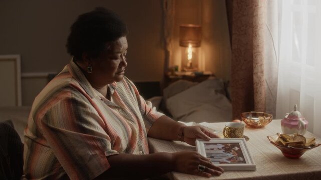 Side view medium shot of senior African American woman sitting alone at table, holding framed photo with melancholic look. Suitable for senior care services, grief counseling, mental health campaigns