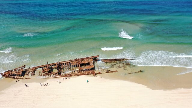 Aerial view of the rusted S.S. Maheno shipwreck resting on the sandy beach with turquoise ocean waves and people walking nearby in K'gari, Queensland, Australia.