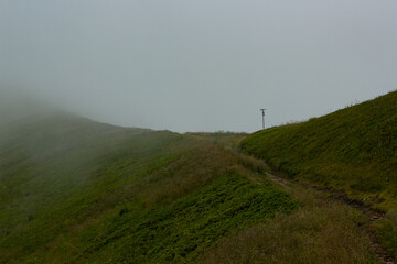 Obraz premium Narrow dirt path winding up a vivid green mountain hill disappearing into low clouds and dense fog. Minimalist moody landscape capturing pure solitude, mystery and the call of wild highland adventure.