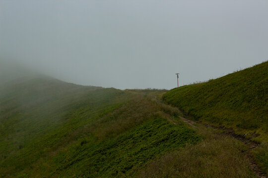Narrow dirt path winding up a vivid green mountain hill disappearing into low clouds and dense fog. Minimalist moody landscape capturing pure solitude, mystery and the call of wild highland adventure.