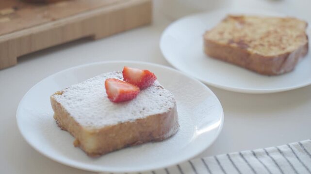 Hand places strawberry slice on French toast dusted with powdered sugar on plate. Concept of homemade breakfast and sweet food preparation