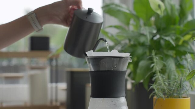 Hand pours water from kettle into paper filter set in coffee dripper on glass server. Concept of pour over coffee preparation and manual brewing