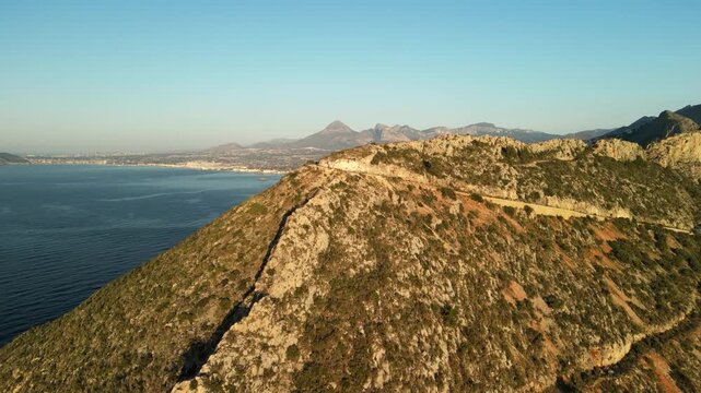 Panoramic aerial view of a rugged Mediterranean coastline with sparse vegetation, leading to a calm sea and distant city against a clear blue sky. Ideal for travel and nature themes.