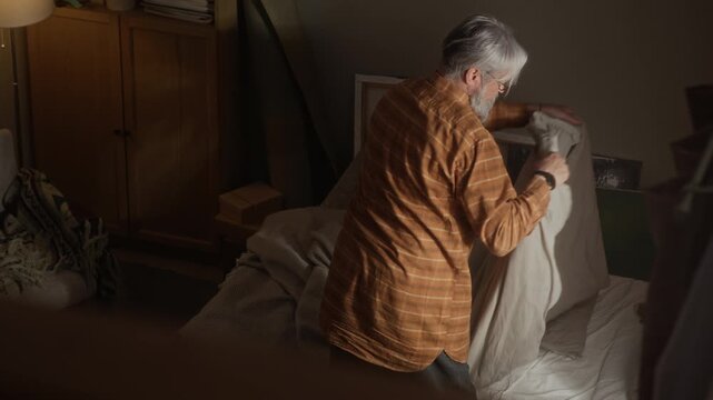 Overhead shot of senior Caucasian man making bed in bedroom, then sitting down looking tired and lonely. Footage is suitable for senior mental health awareness campaigns or retirement home videos