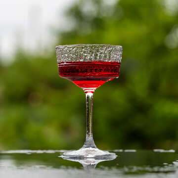 Red cocktail in textured coupe glass on dark reflective surface, outdoors, during the rain. Closeup of refreshing red drink or cocktail in crystal coupe glass against blurred green nature background
