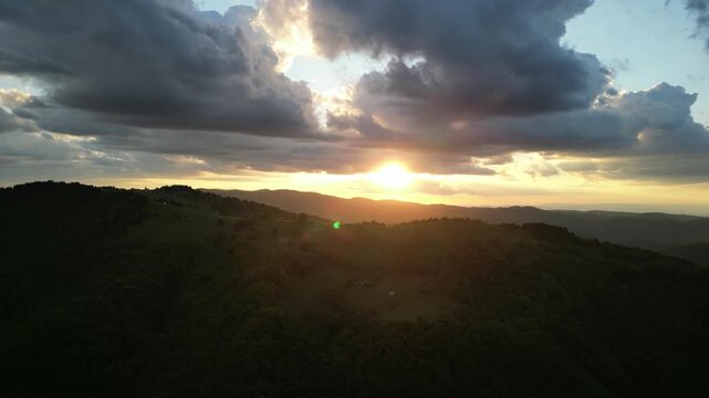 Golden sunset illuminating a serene landscape of rolling green hills and distant forests under a dramatic cloudy sky, creating a peaceful and picturesque natural scene.
