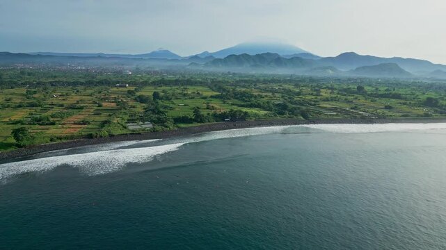 Aerial patchwork rice fields and coastal ponds meeting rocky shore drone mapping shot highlighting irrigation grids, wet paddies, farm tracks and distant volcanic ridge,Bali island